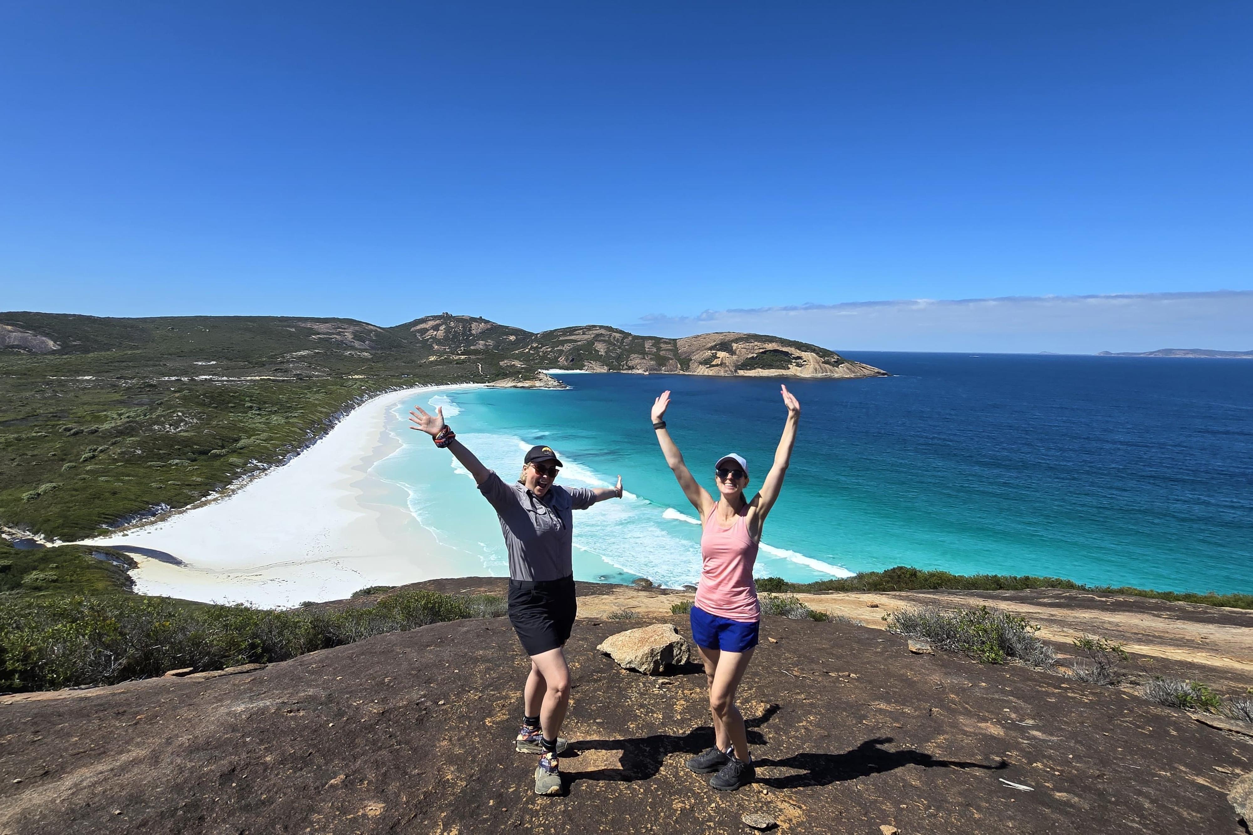 Two people with arms up stretched looking happy on a rock overlooking a white sandy beach and blue ocean. 