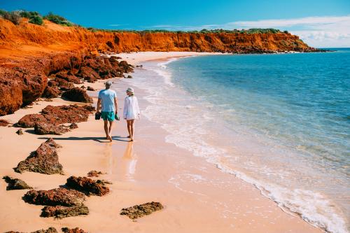 two people walking along sandy beach next to red cliffs and blue clear water 
