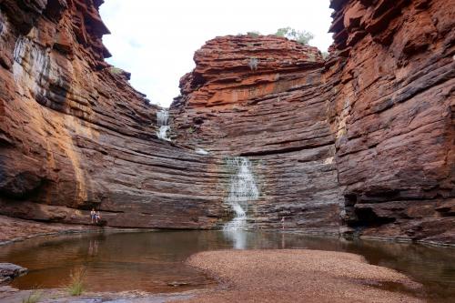 person standing in front of wall of layered dark red rock with a small waterfall