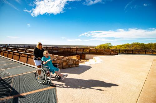 two women one is pushing the other in a wheelchair on a concrete path that leads out to a metal structure overlooking a gorge