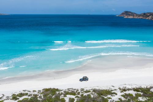 elevated view looking down at a four wheel drive vehicle driving along a white sandy beach with turquoise ocean beyond