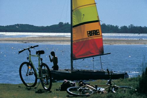 man stabnding in water near catamaran with orange and red sail and two bicycles nearby on grass