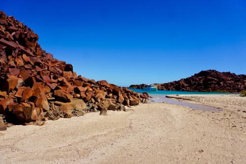 A small bay with red rocks piled high either side and white sandy beach.