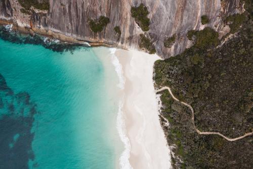 Aerial view of clear blue green water to a white sandy beach. There is a walk trail through the bush to the sand. 