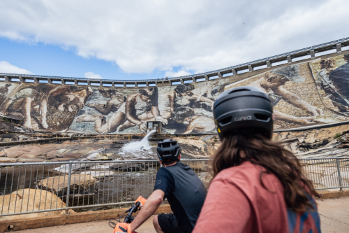 Two bike riders looking at a dam wall painted with a mural. 