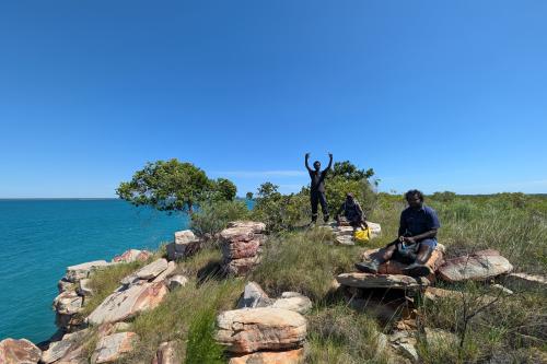 Three people sitting and standing on rocks near the edge of a small cliff on the ocean. 