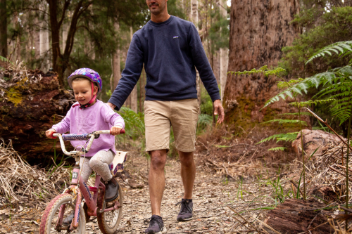 Person walking and helping a child ride a bike on a trail in the forest. 