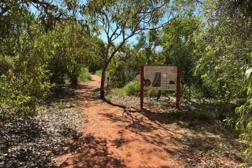 Red dirt trail leading away between trees and bushes. There is a sign showing trail information. 