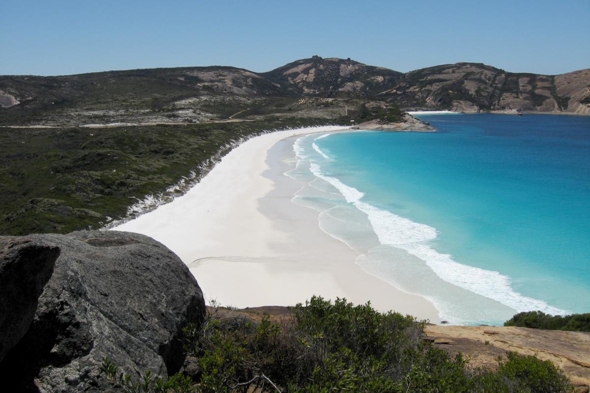 elevated view looking down onto a wide sandy beach with turquoise ocean and tall rock cliffs in the distance