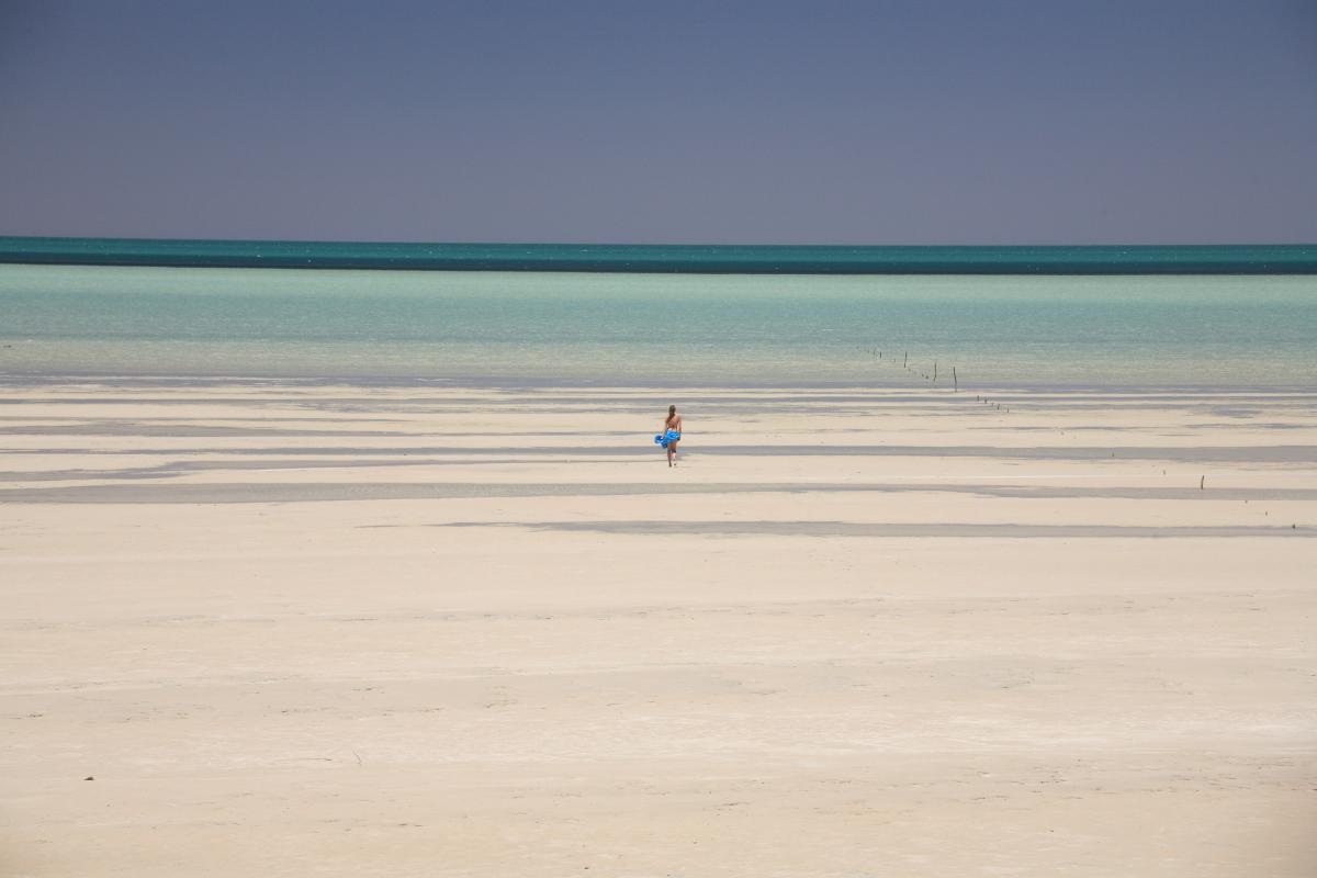 Woman walking on stretch of white sand with clear blue water in the distance