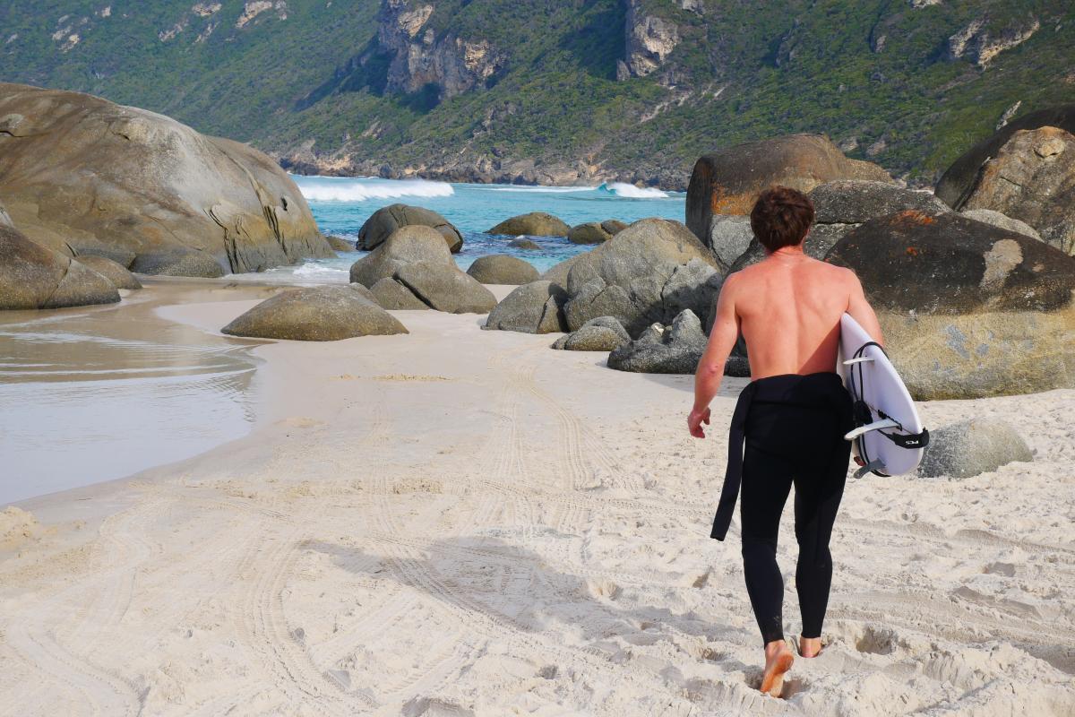 Man in wet suit walking on white sandy beach towards granite boulders and the ocean to go surfing