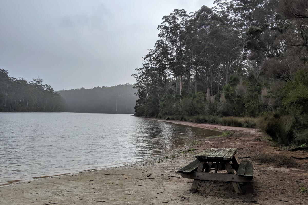 Picnic bench by the water at Big Brook Dam with a rain shower over the forest and water in the distance