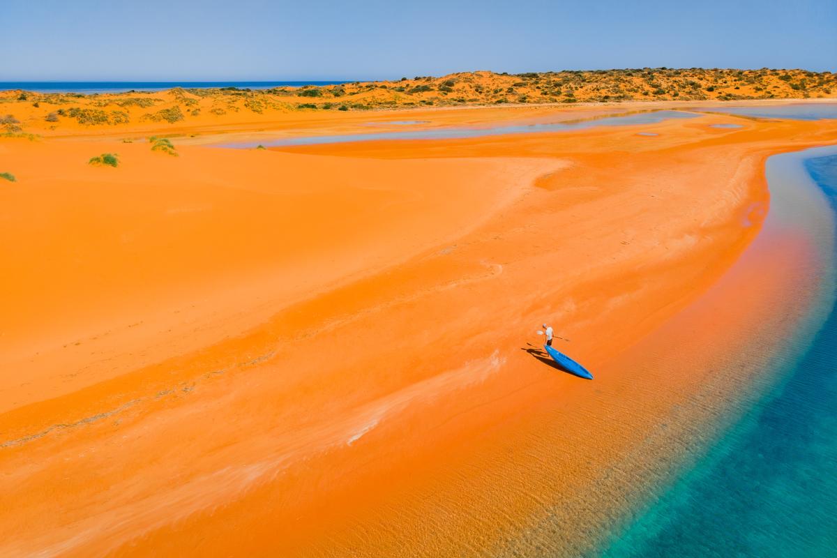 Person dragging kayak on orange sand near clear blue water