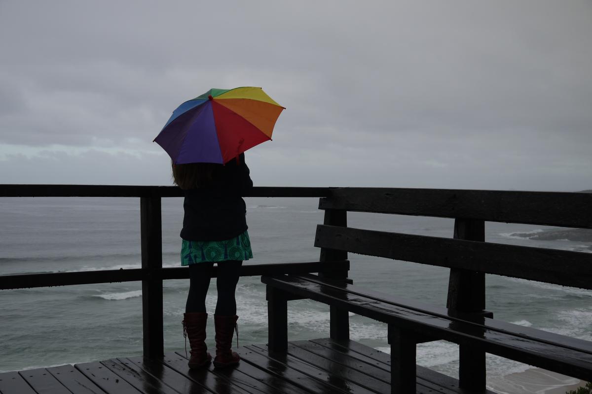 braving the wet weather a lady is standing on the lookout with a multi coloured umbrella and the ocean in the background