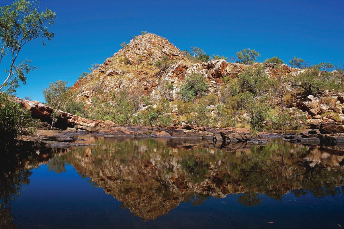 Looking at large hill of red rock next to a waterhole showing the reflection of the rock