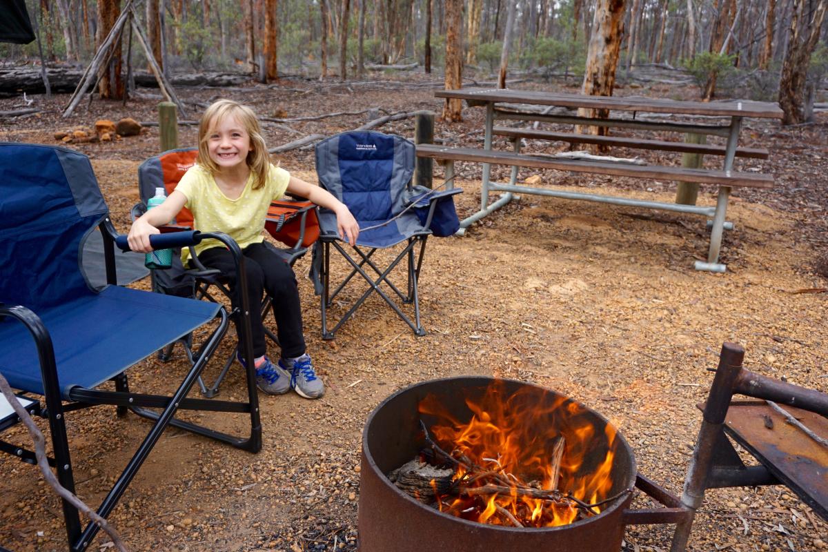 a girl sitting in a camp chair by a drum with a fire in it at a campsite