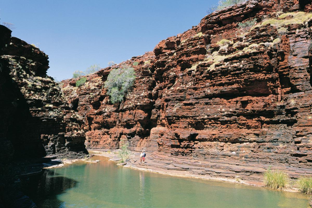 People walking through Kalamina Gorge of layers of red rock and a pool of water