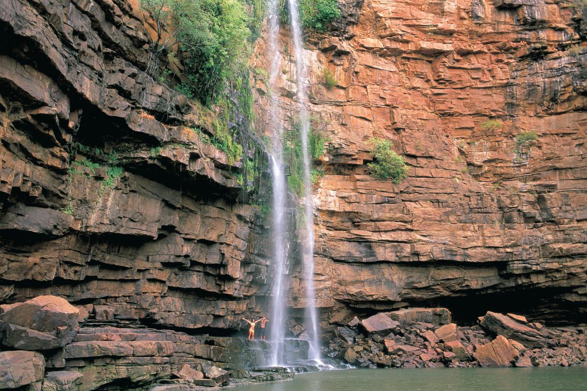 Two people standing on red rock near a long narrow waterfall
