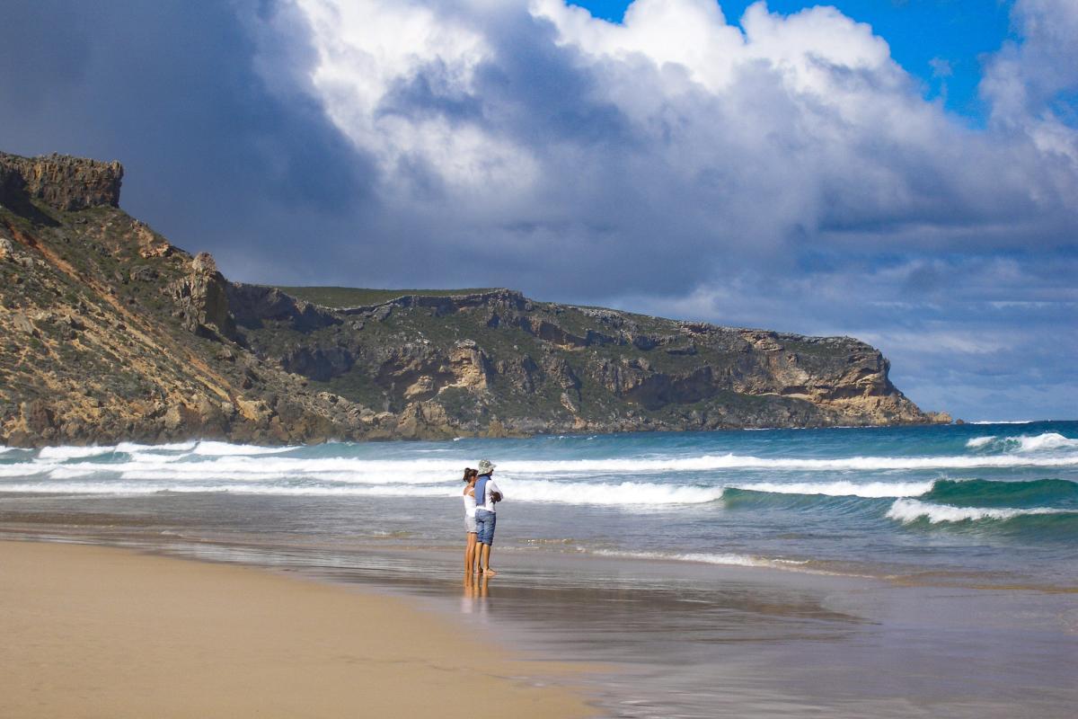 2 people getting their feet wet while standing on the damp sands on a beach and the surf rolling in