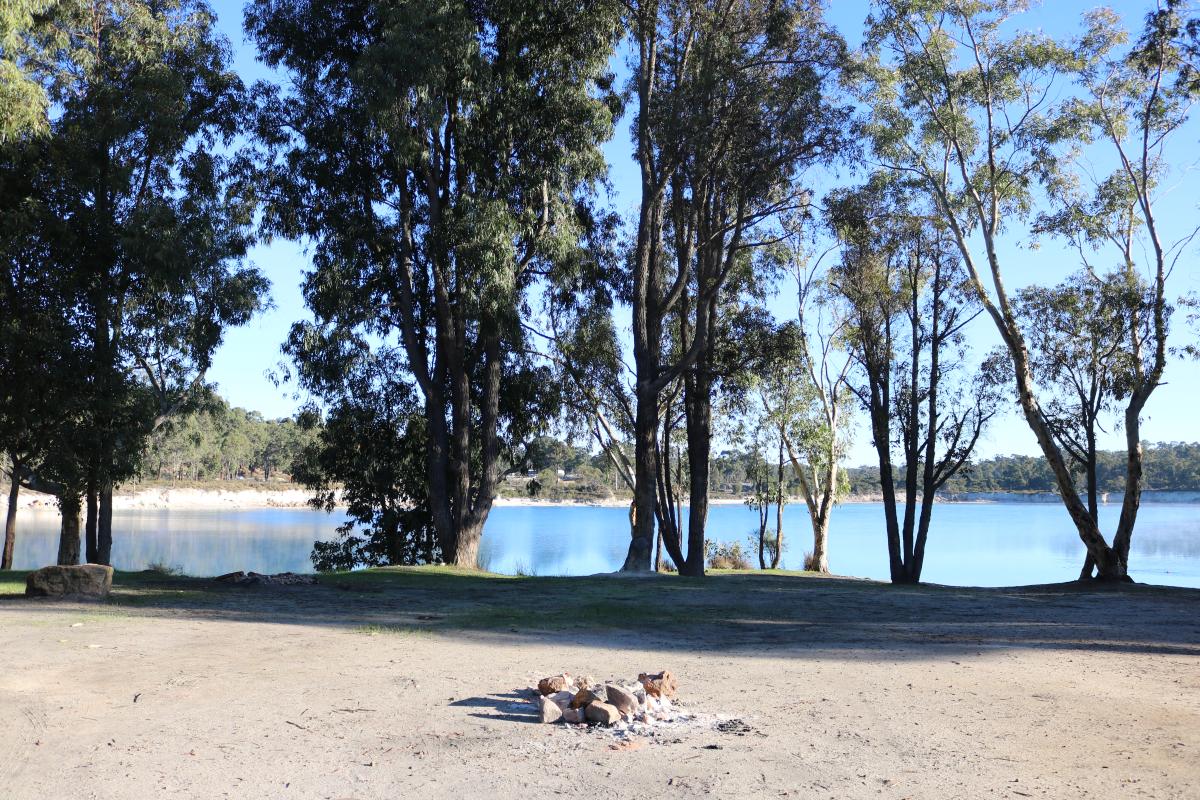 fire ring in a clearing with a row of trees by the dam edge and the blue waters beyond