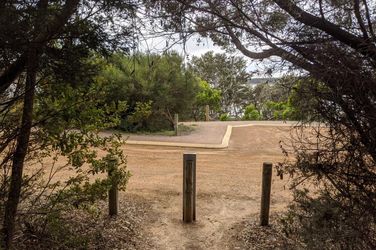 Gravel carpark with low-sloping kerb at the Stokes Inlet day use area viewed from under bushes at the end of the Stokes Heritage Walk Trail