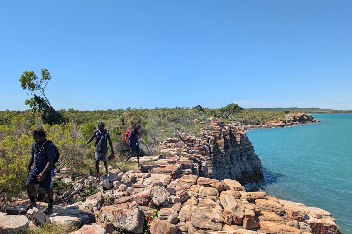 Three people walking on the edge of a small cliff near the ocean. 