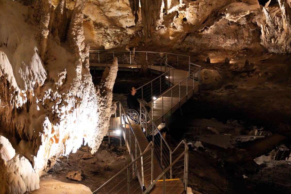 man in wheelchair on boardwalk inside Mammoth Cave, listening to audio tour and looking at cave formations.
