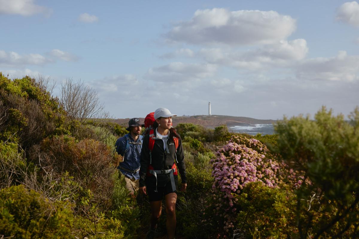 Two hikers walking through shrub with Cape Leeuwin Lighthouse in the distance. 